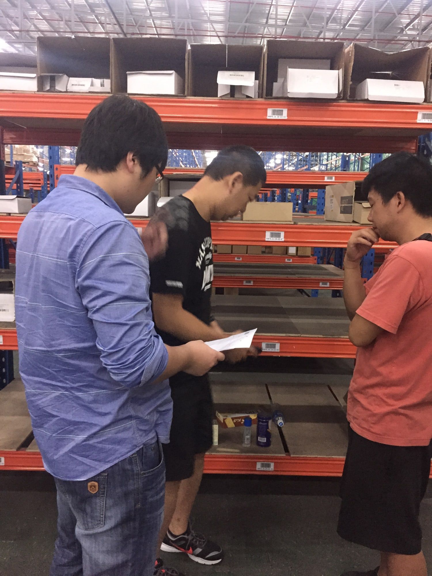 Three warehouse staff read a pick sheet between rows of storage racks.