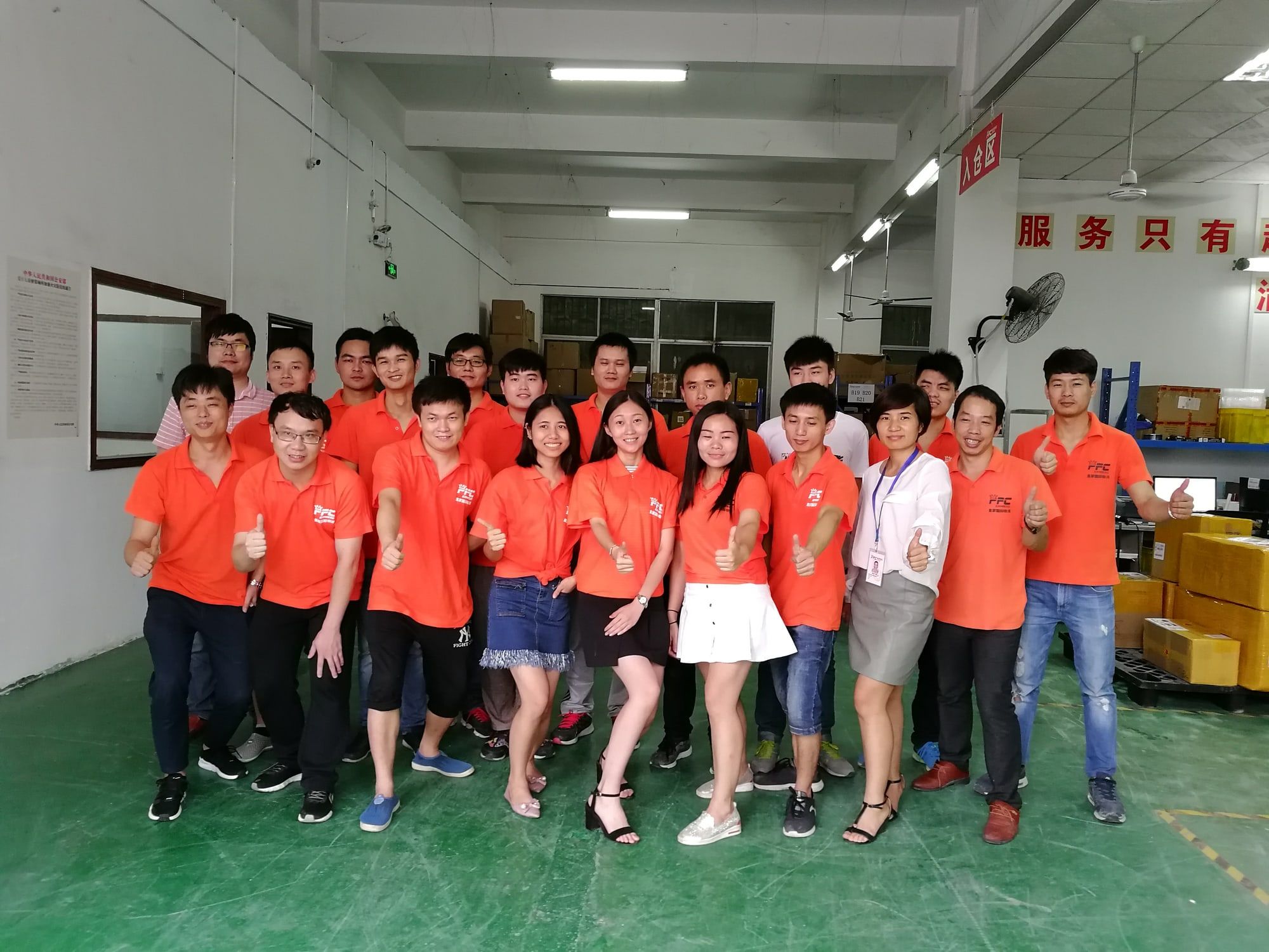 About twenty warehouse staff in orange shirts stand together inside the warehouse.